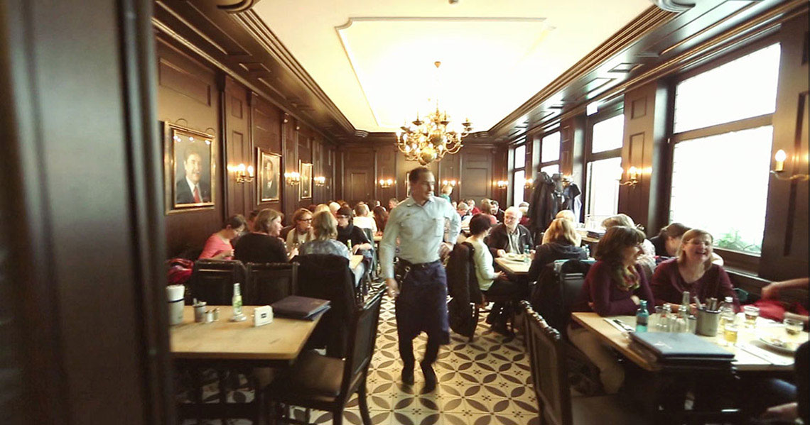 Ein belebtes Restaurant mit einer eleganten dunklen Holzeinrichtung und großen Fenstern. Gäste sitzen an Tischen, ein Kellner geht vorbei.