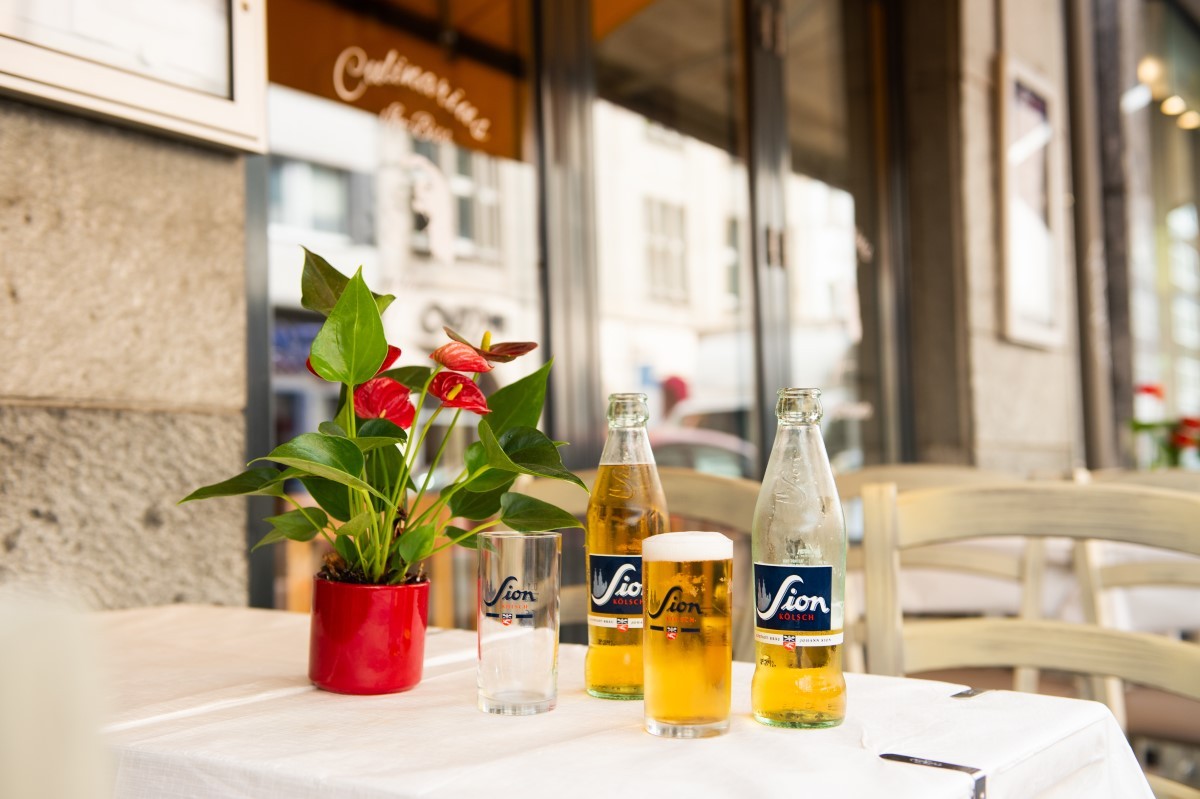 Ein Tisch in einem Straßencafé ist mit zwei Biergläsern und einer leeren Flasche dekoriert. Eine rote Pflanze steht daneben.