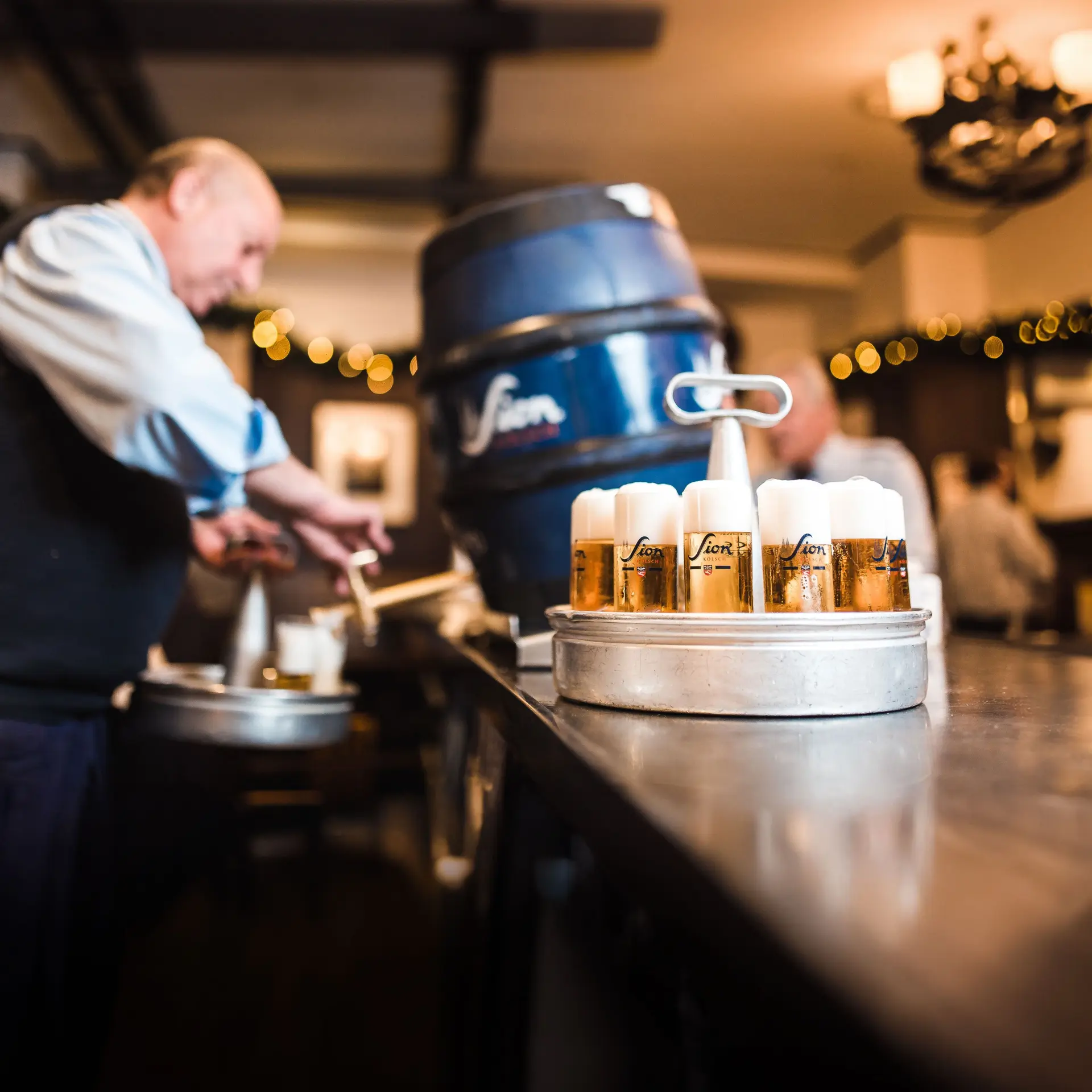Mehrere gefüllte Sion-Biergläser auf einem Tablett an einer Theke. Unscharfer Hintergrund mit Bierfass und Personen.