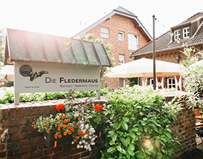 Das Schild "Die Fledermaus" ist auf einem Mauerwerk am Eingang angebracht, umgeben von grünen Pflanzen und Blumen.
