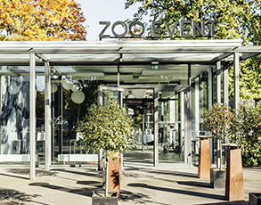 Ein moderner Glas-Eingang eines Zoos umgeben von grünen Bäumen bei Sonnenschein.