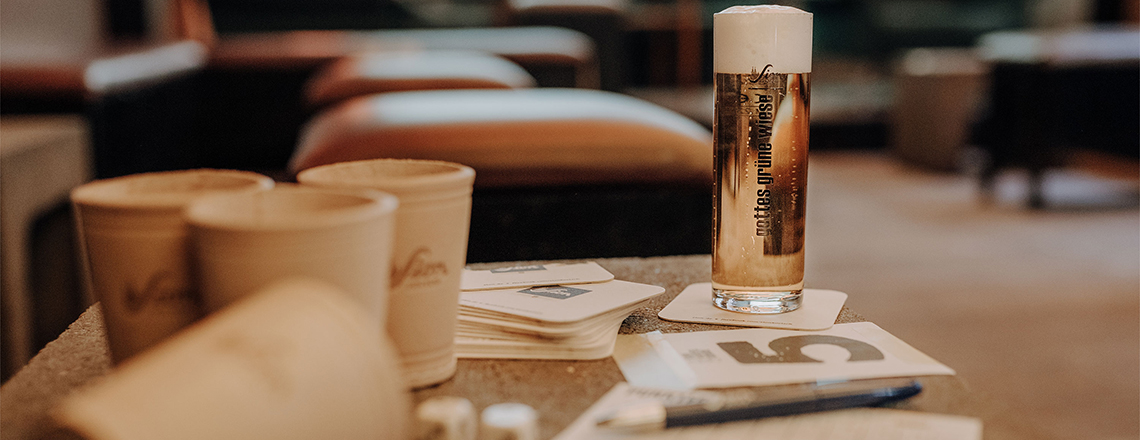 im Vordergrund steht ein Würfelspiel, neben einem Glas Sion Kölsch, dahinter Clubatmosphäre.