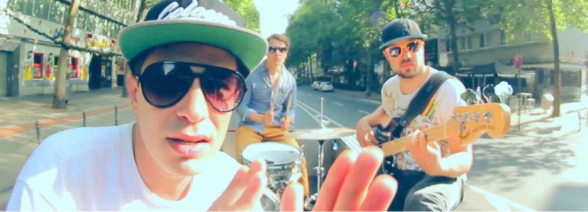 Drei junge Männer spielen Musik auf einer Straße, einer singt in die Kamera.