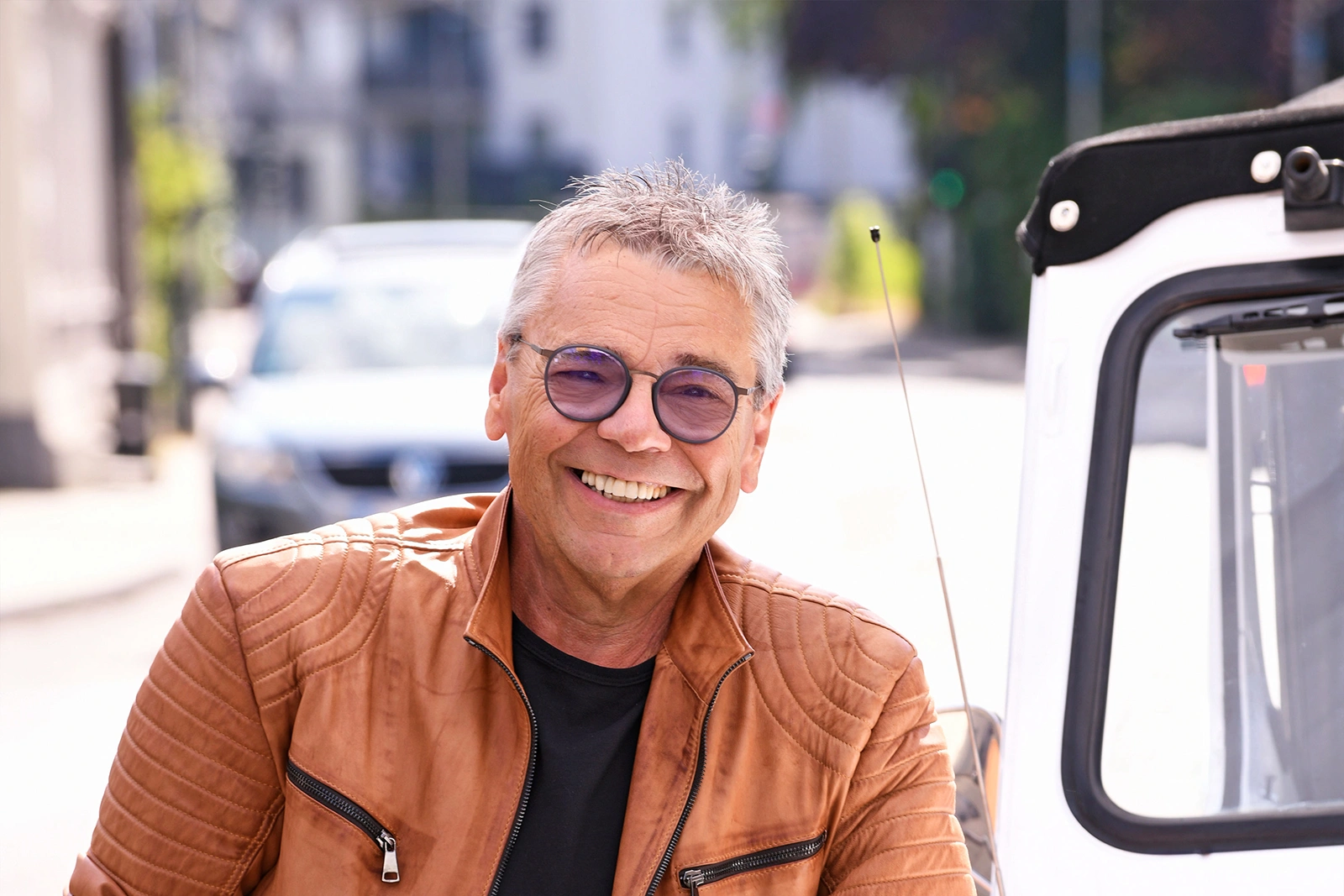 Ein Mann mit grauen Haaren und Brille lächelt in die Kamera. Er trägt eine braune Lederjacke. Im Hintergrund ist eine Straße.