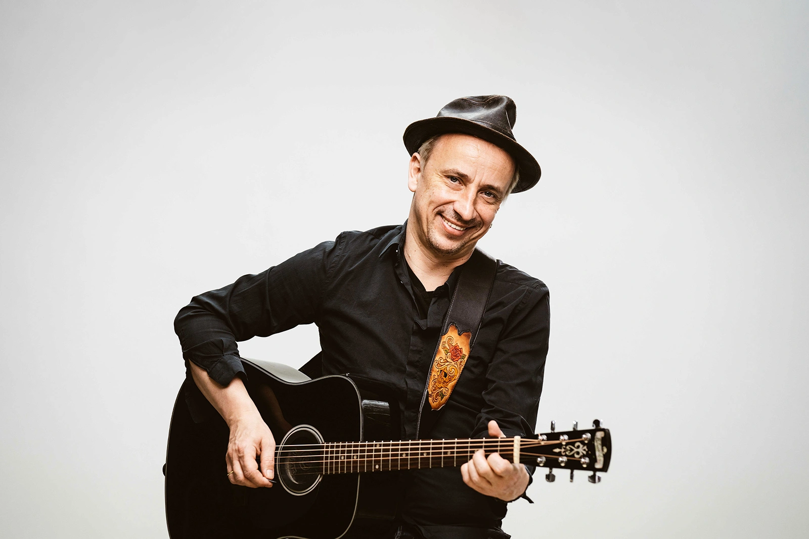 Ein Mann in schwarzem Hemd und Hut spielt eine schwarze Akustikgitarre und lächelt in die Kamera vor einem neutralen Hintergrund.