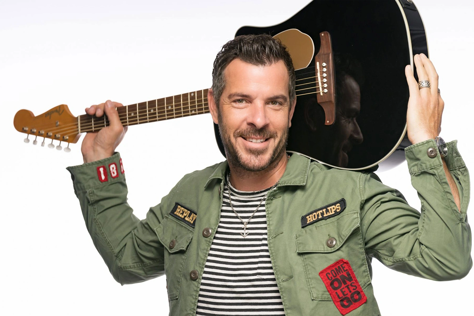 Mann mit Gitarre auf der Schulter, trägt grüne Jacke mit Aufnähern und gestreiftes Shirt. Lächelt direkt in die Kamera.