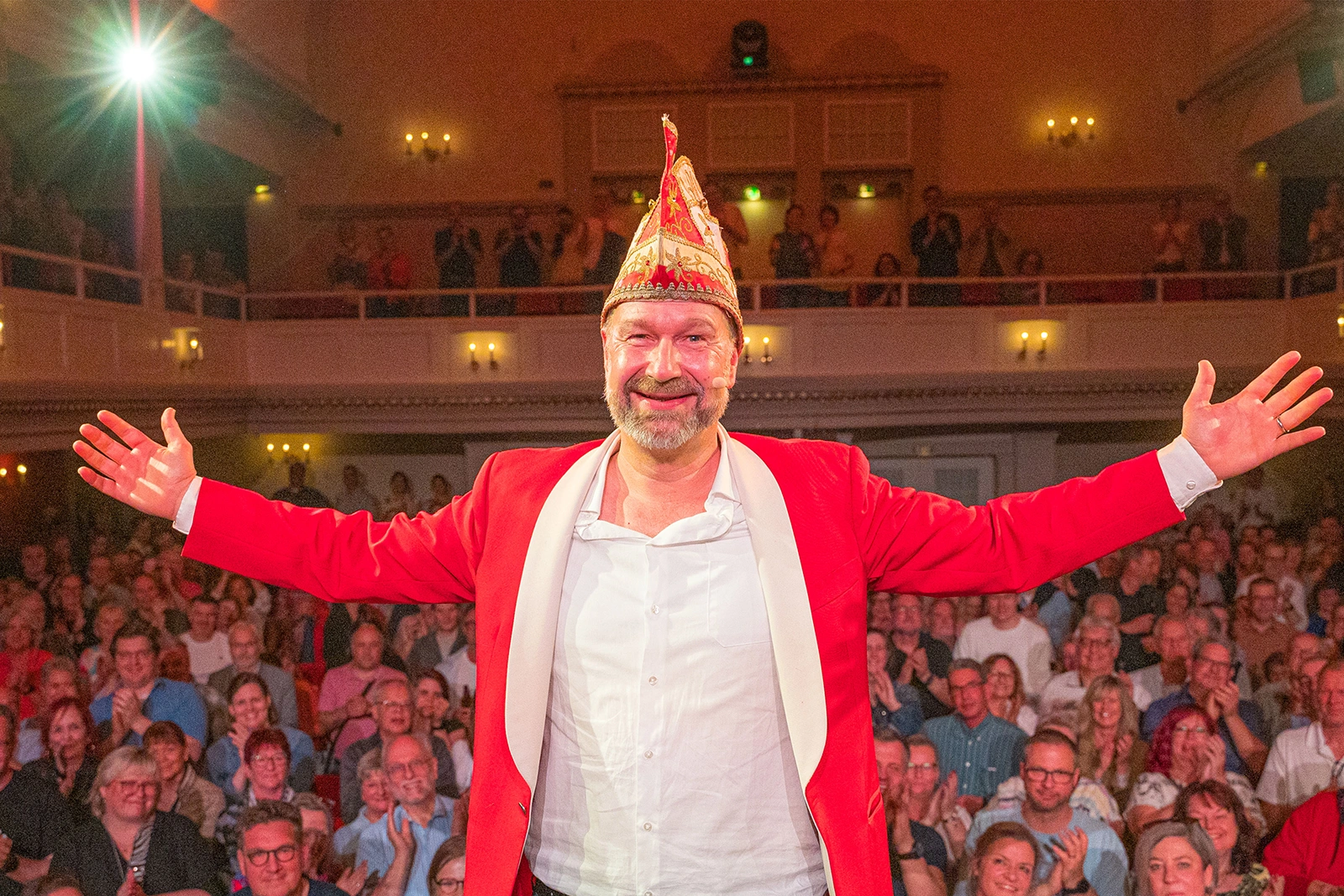 Ein Mann in rotem Anzug und Narrenkappe steht auf einer Bühne vor einem applaudierenden Publikum in einem festlichen Saal.