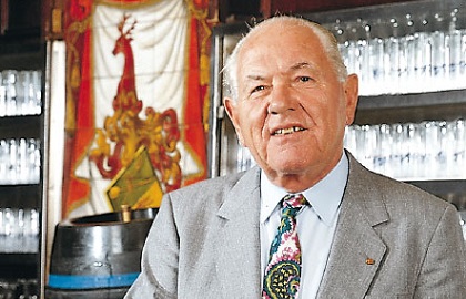 Ein älterer Herr mit grauem Haar und Anzug lächelt vor einem Bar-Hintergrund.