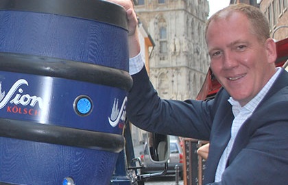 Ein Mann mittleren Alters lächelt, während er eine blaue Bierfass-Säule berührt.