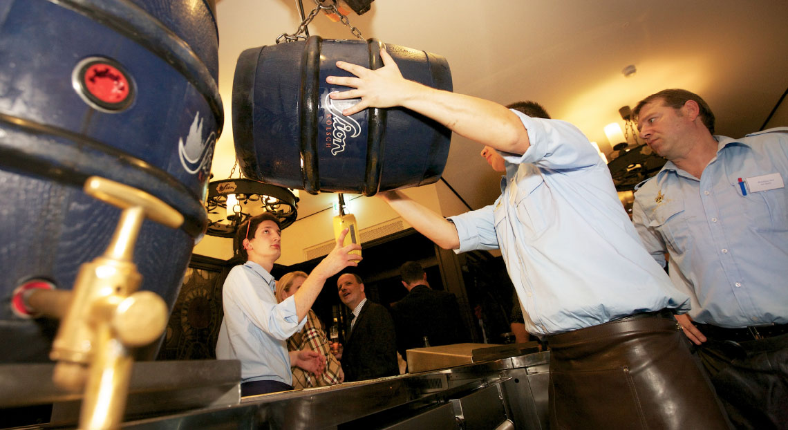 Drei Männer in hellblauen Hemden hängen ein großes Bierfass auf.